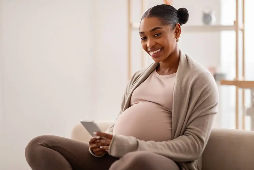 A pregnant woman sitting on a couch smiles while holding a smartphone. She wears a beige cardigan over a light shirt and has her hair in a bun. The background is softly lit with shelves and a vase visible.