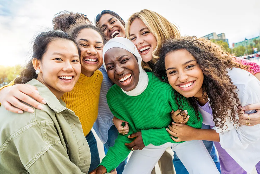 A group of six diverse young women standing close together outdoors, smiling and laughing as they pose for a photo, showing joy and friendship.